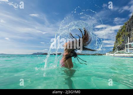 Philippines, Palawan, archipel de Calamianes, zone biotique naturelle de l'île de Coron, île de Coron, plage de Banul, jeune philippine en bikini, se tenant la tête hors de l'eau de mer turquoise et soufflant ses longs cheveux sombres dans une grande éclaboussure Banque D'Images