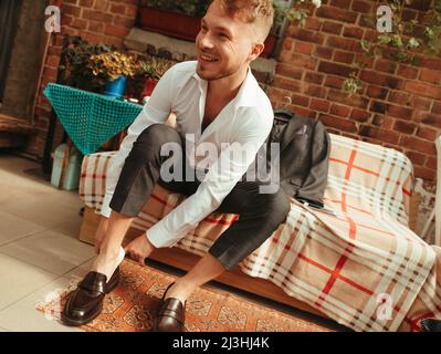 L'homme a ses chaussures élégantes dans sa chambre. Jeune homme d'affaires élégant assis sur le lit dans la chambre d'hôtel et portant ses chaussures élégantes. Effet grain Banque D'Images