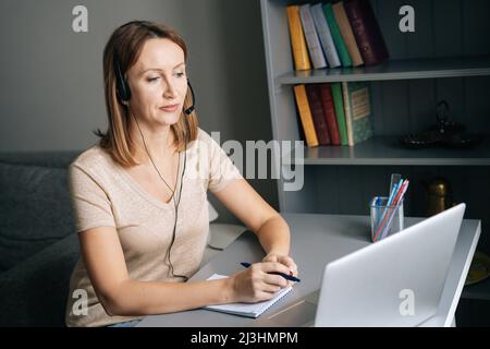 Une jolie femme concentrée dans des écouteurs étudiant en ligne sur un ordinateur portable, prenant des notes, travaillant à distance sur un ordinateur de bureau à domicile. Banque D'Images