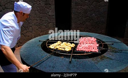 Îles Canaries, Lanzarote, île volcanique, parc national de Timanfaya, paysages volcaniques, gril alimenté par l'énergie géothermique, sur lui des saucisses et d'autres aliments grillés, à côté du gril tient un cuisinier dans les vêtements de travail blancs et avec un capuchon blanc et déplace la grille Banque D'Images
