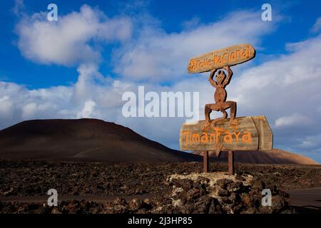 Îles Canaries, Lanzarote, île volcanique, parc national de Timanfaya, paysages volcaniques, route d'entrée au parc national, panneau d'entrée en bois, figure du monument de Lanzarote, le diable de feu est sur le panneau 'Timanfaya', avec ses mains levées il tient le panneau avec l'inscription 'Parque Nacional ', ciel bleu avec des nuages blancs, la roche de lave se trouve autour du panneau Banque D'Images