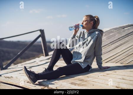 Femme sportive se reposant après l'exercice et l'eau potable tout en étant assise sur le vieux pont dans la nature. Banque D'Images