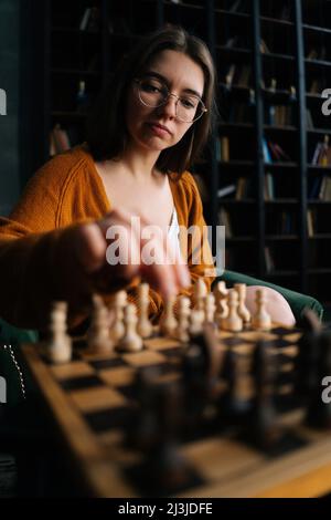 Portrait vertical en gros plan de la jeune femme dans des lunettes élégantes faisant des échecs se déplacer assis sur un fauteuil dans la pièce sombre, foyer sélectif. Banque D'Images