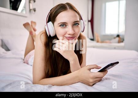 Une femme heureuse tient le téléphone dans son casque pendant qu'elle est dans son lit. Banque D'Images