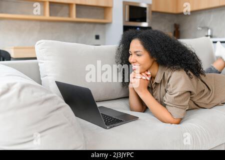 Jeune femme indépendante ou étudiante intelligente et positive, posée sur le canapé avec un ordinateur portable, étudiant à partir d'un séminaire en ligne ou d'un cours à la maison, visiophonie et conversation Banque D'Images