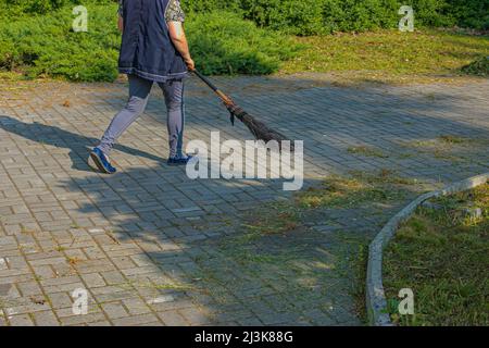 Une femme balaie les feuilles mortes avec un balai dans la rue. Le service municipal surveille la propreté de la ville. Banque D'Images