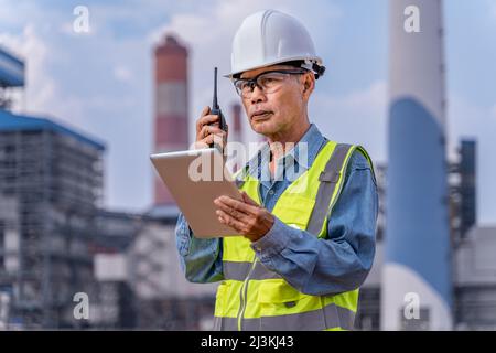 Un ingénieur superviseur senior dans ses chiffons de travail avec un casque blanc, des lunettes de sécurité utilisant sa radio talkie-walkie pour communiquer avec son équipe pendant la période de l'Hol Banque D'Images