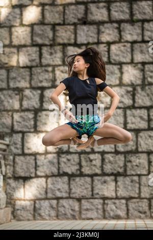 Fille dansant avec des chaussures de ballet et dans un air de pose contre un mur de pierre dans un parc public; Hong Kong, Chine Banque D'Images