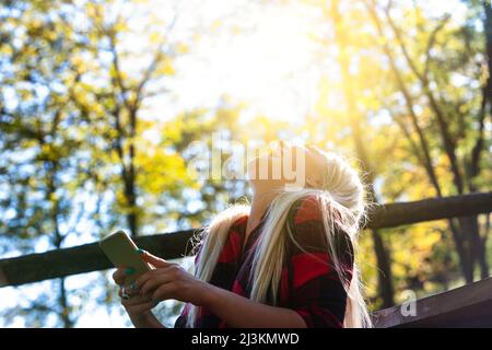 Une femme rieuse, éclairée par la lueur chaleureuse du soleil qui éclate à travers des arbres verts verdoyants, jette sa tête en riant joyeusement Banque D'Images