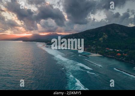 Vue aérienne des quais et des brise-lames le long de la rive de la plage de Candidasa avec la lumière du soleil qui traverse les sommets des montagnes sous un ciel gris nuageux... Banque D'Images