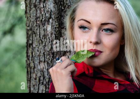 Jeune femme pensive mâchant sur une feuille verte comme elle se détend sur le tronc d'un arbre dans la forêt Banque D'Images