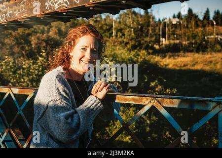 Portrait extérieur d'une femme mûre aux cheveux bouclés rouges debout sur un pont dans un parc en automne; Edmonton, Alberta, Canada Banque D'Images