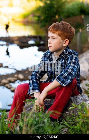 Portrait extérieur d'un garçon aux cheveux rouges assis à côté d'un ruisseau; Edmonton, Alberta, Canada Banque D'Images