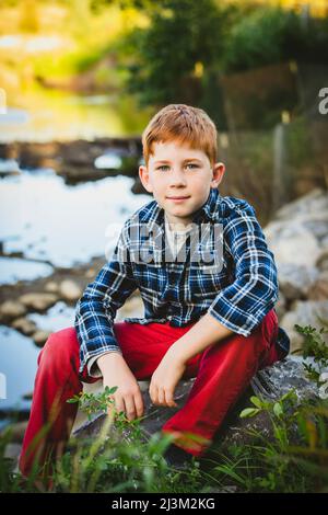 Portrait extérieur d'un garçon aux cheveux rouges assis à côté d'un ruisseau; Edmonton, Alberta, Canada Banque D'Images