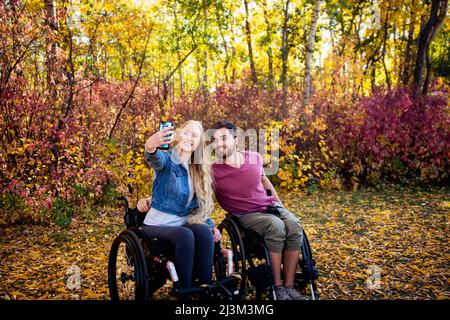 Un jeune homme et une jeune femme paraplégique en fauteuil roulant prenant un autoportrait avec un smartphone dans un parc lors d'une belle journée d'automne Banque D'Images