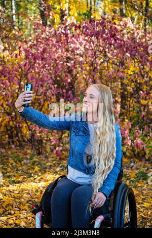 Une jeune femme paraplégique dans son fauteuil roulant qui prend un autoportrait avec son téléphone intelligent dans un parc lors d'une belle journée d'automne; Edmonton, Alberta, Canada Banque D'Images