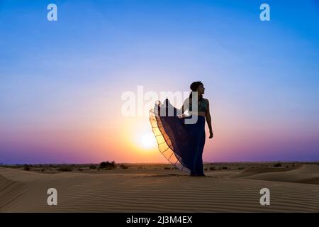 Femme à longue jupe rétro-éclairée par le coucher du soleil sur les sables du désert ; Abu Dhabi, Émirats arabes Unis Banque D'Images