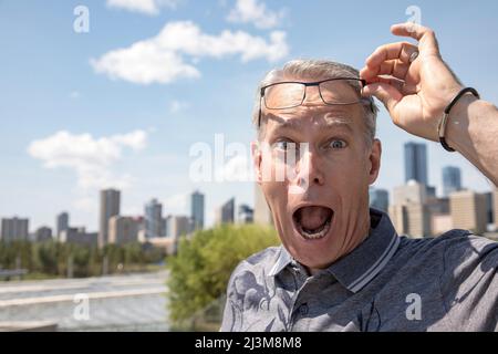 Homme âgé debout à l'extérieur, levant ses lunettes pour regarder la caméra avec une expression surprise sur son visage; Edmonton, Alberta, Canada Banque D'Images