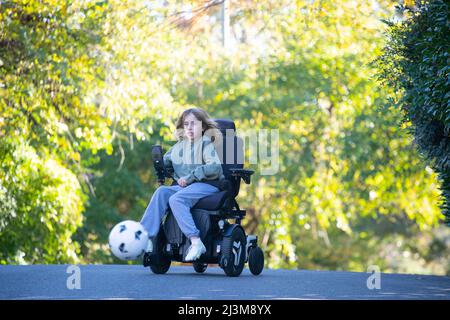 Une jeune fille de 11 ans atteinte de dystrophie musculaire congénitale d'Ullrich joue au football dans son fauteuil roulant près de sa maison Banque D'Images