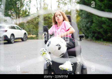 Une jeune fille de 11 ans atteinte de dystrophie musculaire congénitale d'Ullrich joue au football dans son fauteuil roulant près de sa maison Banque D'Images