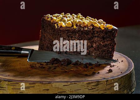 Un morceau coupé de gâteau au chocolat recouvert de noix sur un tronc d'arbre coupé avec un couteau; Studio Banque D'Images