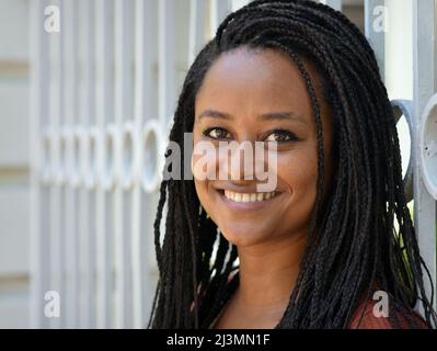 Gaie positive belle jeune femme africaine sombre à la peau avec de longs et stylés braides Afro sourires pour le spectateur devant une clôture en fer forgé. Banque D'Images