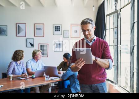 Le Digital Manager fait tout. Photo d'un homme mûr utilisant une tablette numérique avec son équipe dans le fond d'un bureau moderne. Banque D'Images