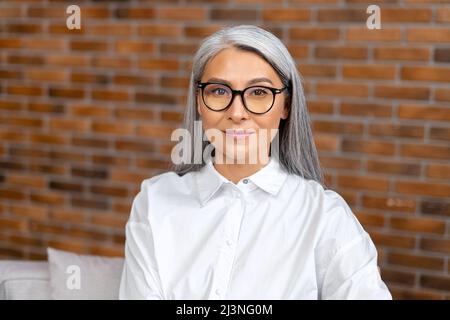Contemporaine satisfaite, la femme âgée porte des lunettes aime le matin tout en étant assise sur le fauteuil au bureau. Calme et insouciante de la retraite dame rêve, regarde la caméra et sourire Banque D'Images