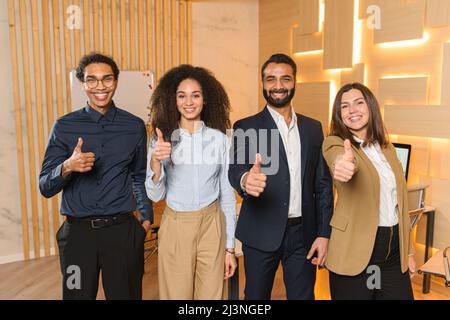 Une équipe joyeuse et réussie de jeunes professionnels portant des vêtements élégants et décontractés debout au bureau et montrant de grands doigts. Atmosphère informelle en équipe multiraciale Banque D'Images
