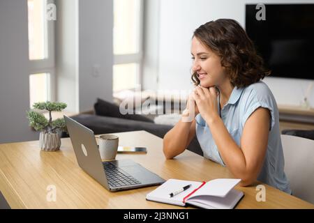 Charmante femme caucasienne regardant des webinaires en ligne sur l'ordinateur portable assis à la table à la maison, prenant des notes, e-learning. Femme d'affaires senior recherchant, analysant les tâches travaille dans le bureau à domicile Banque D'Images