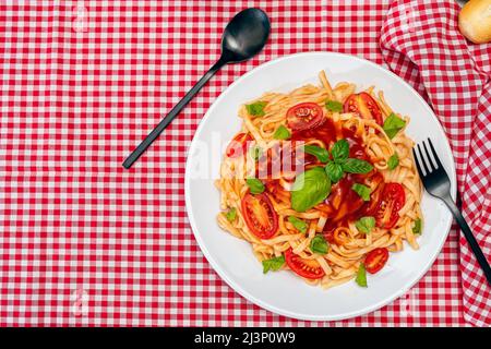 Vue de dessus des pâtes spaghetti avec une délicieuse sauce tomate maison avec des feuilles de basilic faites maison servies sur une assiette blanche sur une nappe à carreaux rouges. C Banque D'Images