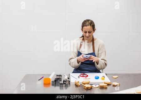 Femme pâtissière utilisant son téléphone portable tout en travaillant dans la cuisine de la pâtisserie. Banque D'Images