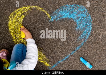 Arrêter la guerre en Ukraine. Une petite fille dessine un coeur en jaune et bleu avec de la craie sur le trottoir comme le drapeau ukrainien. Photo de haute qualité Banque D'Images