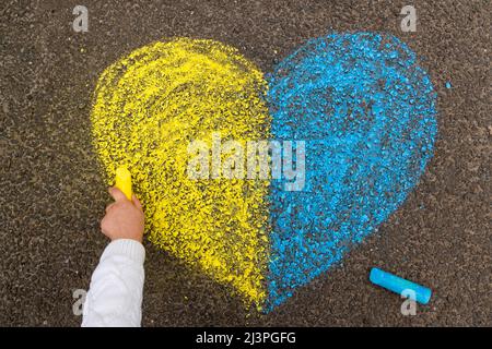 Arrêter la guerre en Ukraine. Une petite fille dessine un coeur en jaune et bleu avec de la craie sur le trottoir comme le drapeau ukrainien. Photo de haute qualité Banque D'Images