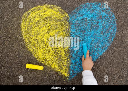 Arrêter la guerre en Ukraine. Une petite fille dessine un coeur en jaune et bleu avec de la craie sur le trottoir comme le drapeau ukrainien. Photo de haute qualité Banque D'Images