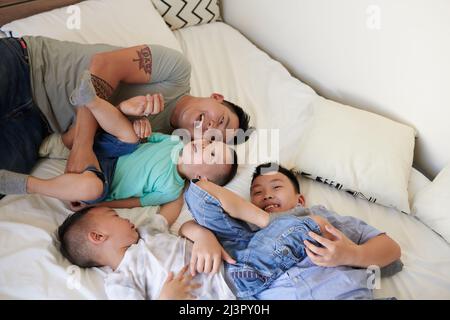 Un père joyeux et ses trois fils se détendent au lit lorsqu'ils passent un week-end ensemble à la maison Banque D'Images