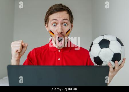 Jeune homme enthousiaste soutien de fan Allemagne équipe nationale de football tenir à la main ballon de football regarder la télévision en direct sur ordinateur portable. Banque D'Images