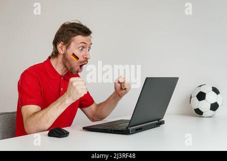 Jeune homme enthousiaste fan de maillot rouge soutien de l'équipe nationale de football allemande tenir à la main ballon de football regarder la télévision en direct sur ordinateur portable. Banque D'Images