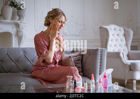 Portrait belle femme d'âge moyen regardant dans le miroir assis sur un canapé confortable à la maison, jolie femme touchant la peau de cou posant pour le phot, la beauté naturelle et les soins de la peau assis sur le canapé en peignoir rose Banque D'Images