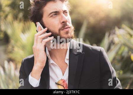 Beau barbu jeune homme en costume parlant au téléphone à l'extérieur. Banque D'Images