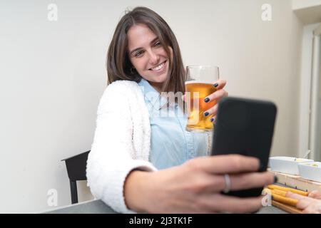Une jeune femme boit de la bière lors d'un appel vidéo avec son ami ou ses amis. Elle est assise à une table dans la cuisine à la maison. Banque D'Images