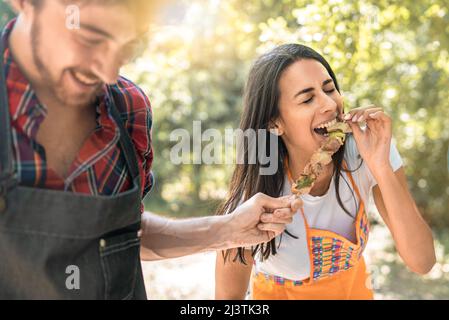 une jeune femme rit alors qu'elle mange des brochettes de viande des mains de son ami lors d'un voyage à la campagne. Concept de style de vie des jeunes adultes ayant le fu Banque D'Images