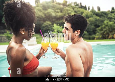Couple biracial de jeunes gens assis au bord de la piscine dans la nature regardant les uns dans les autres les yeux et toaster avec de longs cocktails alcoolisés dans le summ Banque D'Images