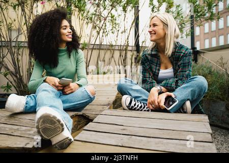 De belles jeunes filles multiraciales qui se reposent sur la terrasse avec leur smartphone. Conversation émotionnelle entre amies multiculturelles. Banque D'Images