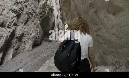 Vue arrière d'une jeune femme marchant dans une gorge entre des rochers avec un téléphone dans ses mains. Action. Femme voyageur photographiée de belles pierres naturelles Banque D'Images