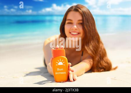 Portrait d'une femme magnifique souriant tout en se relaxant sur la plage et en tenant une bouteille de crème solaire Banque D'Images