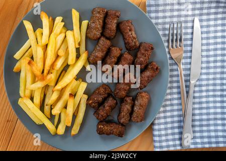 CEVAPi, plat traditionnel de viande hachée des balkans, servi avec des frites dans une assiette bleu marine sur une table en bois Banque D'Images