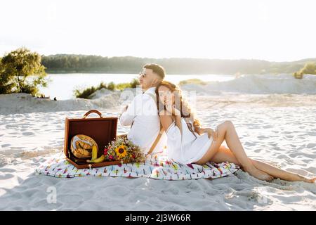 Pique-nique au coucher du soleil. Couple aimant assis sur un tapis au bord du lac Banque D'Images