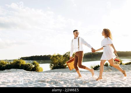 Couple heureux courant sur le sable, coucher de soleil, couple élégant avec une valise et des fleurs dans un panier. Banque D'Images