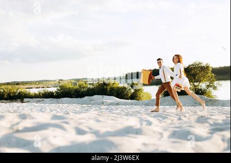 Couple heureux courant sur le sable, coucher de soleil, couple élégant avec une valise et des fleurs dans un panier. Banque D'Images